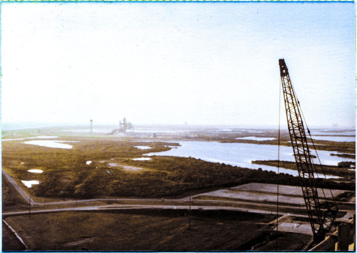 Image 042. In the golden light of a Florida Early Morning, the Space Shuttle Columbia sits poised in its launch position, supported by the Mobile Launch Platform which spans the Flame Trench at distant Launch Complex 39-A, Kennedy Space Center, Florida. You are seeing Columbia, prior to its second-ever mission, from a vantage point up on the Fixed Service Structure, high above the surrounding wilderness, at Pad B, which, at the time this photograph was taken, was still under construction, and had yet to host its own Shuttle launches. To the right, you can see the jib of Wilhoit Steel Erectors big Manitowoc crane, which the Union Ironworkers of Local 808 were using to erect the Rotating Service Structure, which is just out of vie To w, below and to the right. Photograph by James MacLaren.